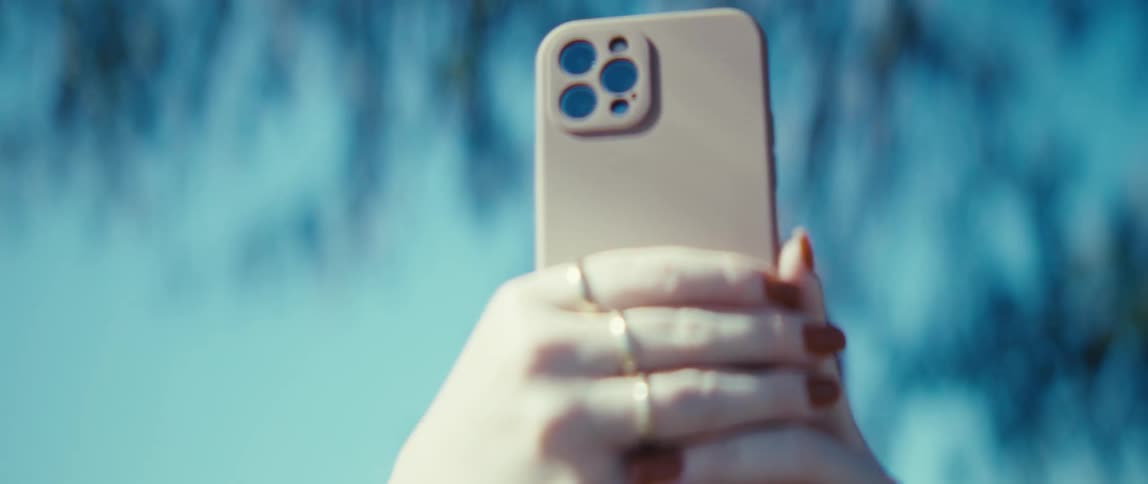 Person wearing glasses holds phone with beige case against blue sky and blurred branches