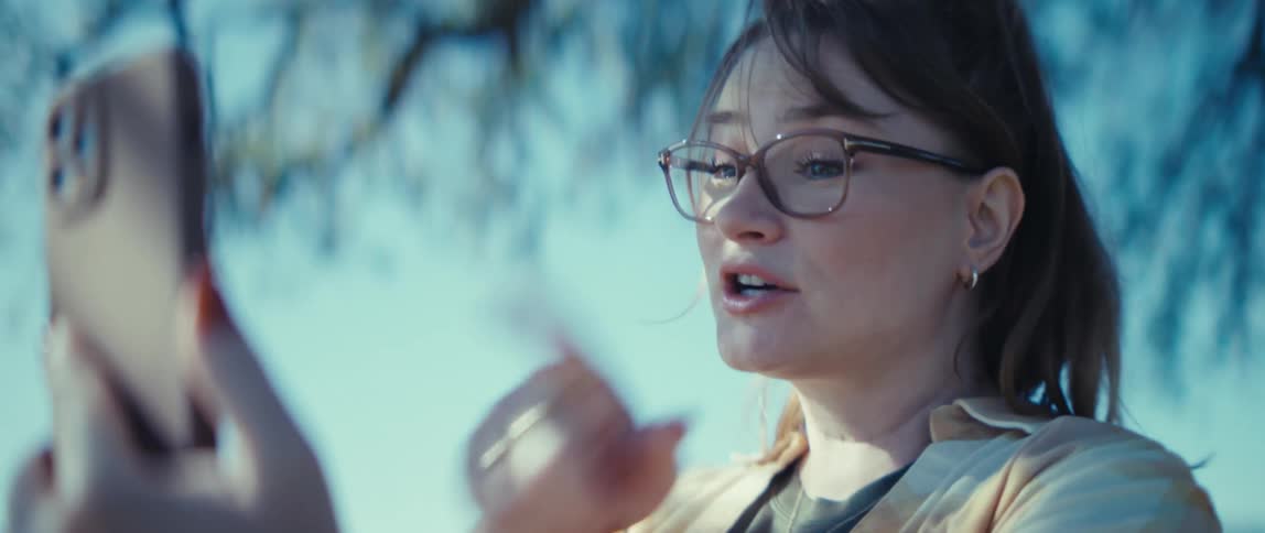 Woman wearing glasses and light shirt holds phone with left hand under blue sky and blurred branches