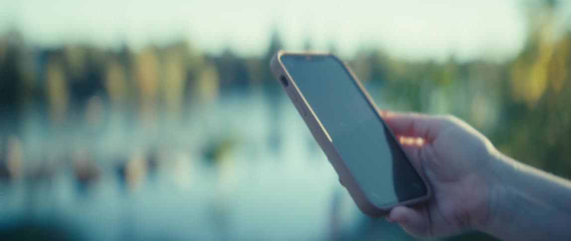 A close view shows a hand holding a modern smartphone with a pink case with the reflection of water and blurred green trees in the bright sunlight in the background
