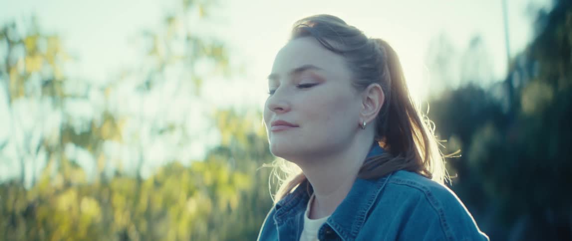 The subject is a young woman with reddishbrown hair tied back in a ponytail wearing a denim jacket over a white shirtshe is shown outdoors looking offcamera to the side with soft natural lighting creating a bright somewhat hazy background of gre