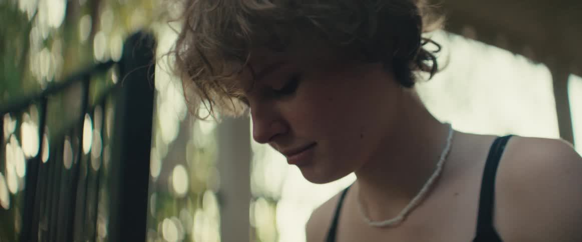 The video is a closeup of a young person with curly short hair and a beaded necklace looking down with a subtle smilethey are outdoors in a brightly lit natural setting with diffused sunlight filtering through the background foliage