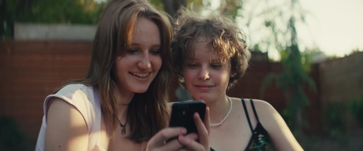 Two girls stand close together and look down at a mobile phone screen held by one of them both girls smile and react to the screen content and the surrounding area is blurred and lit by warm lateday sunlight