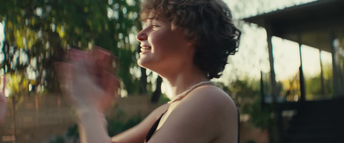 Two smiling young women face each other and clap their hands excitedly outdoors with bright sunlight and green trees creating lens flare in a celebratory atmosphere