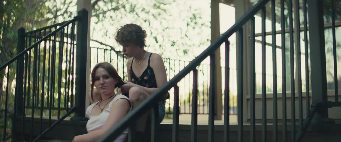 Two young women sit on outdoor stairs with a black metal railing one person sits higher and braids the other persons hair and they are surrounded by trees and bright daylight