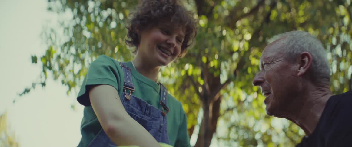 Young person in green shirt and denim overalls smiles beside elderly woman with gray hair in sunlit natural setting
