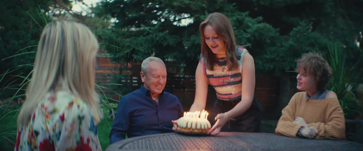 The video shows an outdoor birthday celebration where a woman lights the candles on a bundt cake topped with whipped cream or frostingthe woman holds the cake over a table while a seated group including an older man in a navy shirt smiles and