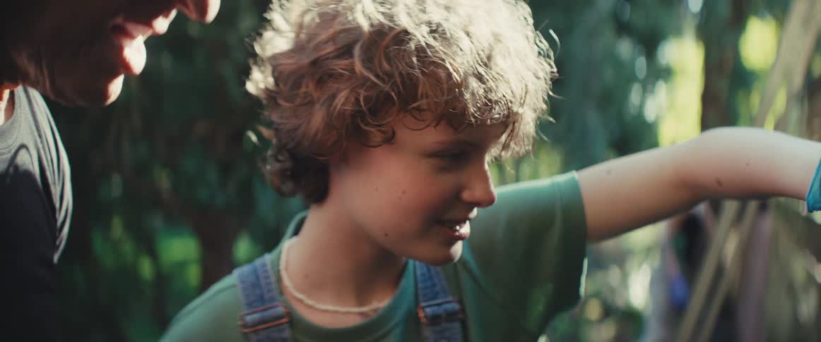 Boy is outdoors next to a wooden fence accompanied by an adult