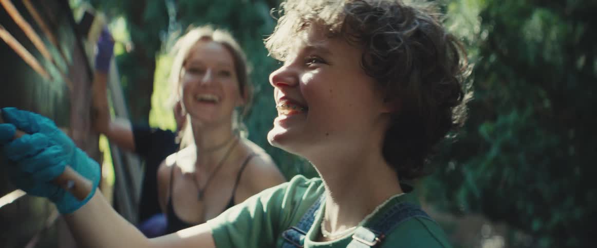 Two teenager girls and an adult man talking and laughing while painting a wooden fence outdoors during day time
