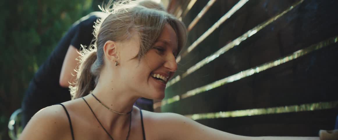 The video is a brightly lit medium shot of a woman smiling while painting a dark wooden fence outdoorsshe is wearing blue gloves and using a brush suggesting she is enjoying a communal activity or an outdoor project