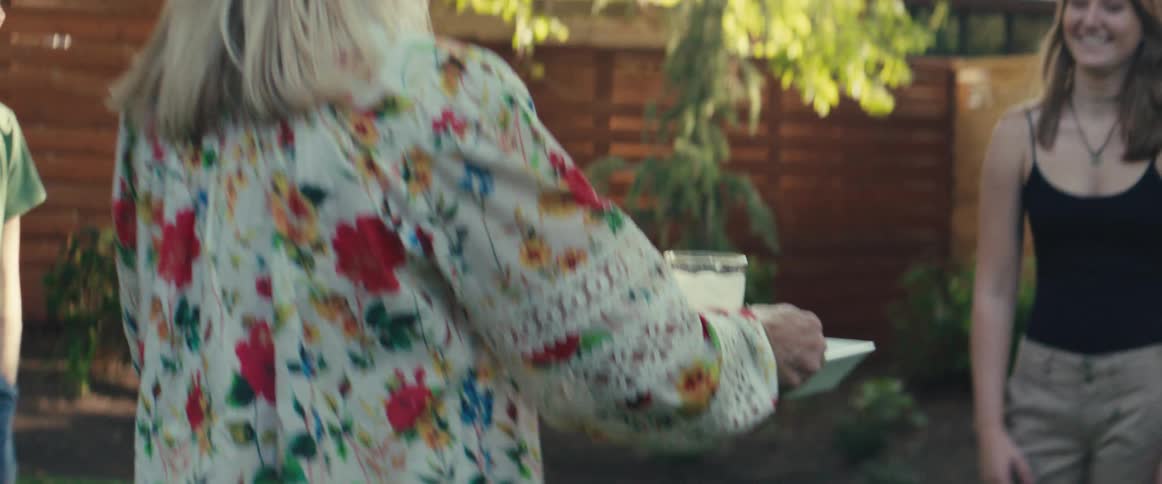 Person in patterned shirt holds tray with pitcher and bowl in outdoor area with wooden fence trees and other people in background