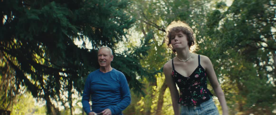 A young woman with curly hair and a floral tank top walks and then claps happily while smiling at an older man next to her the two are outdoors in a brightly sunlit park or wooded area with large evergreen trees in the background