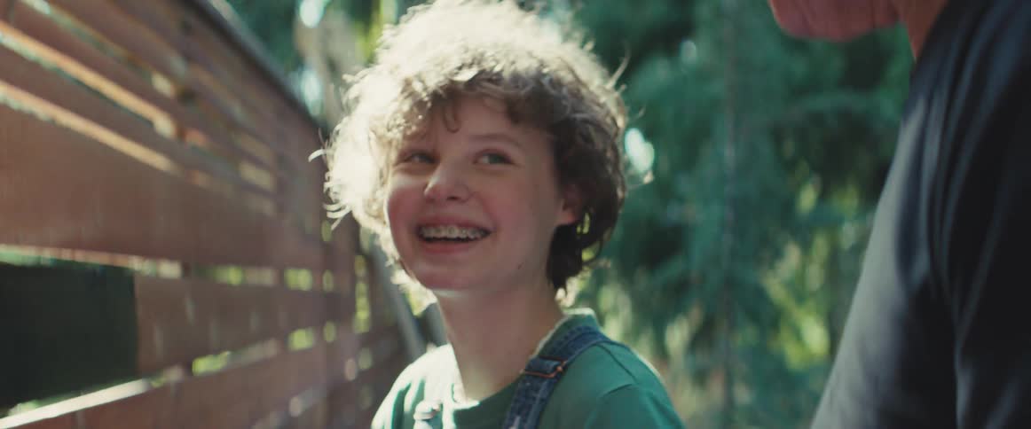 Boy smiles in outdoor area with wooden fence and greenery in the background
