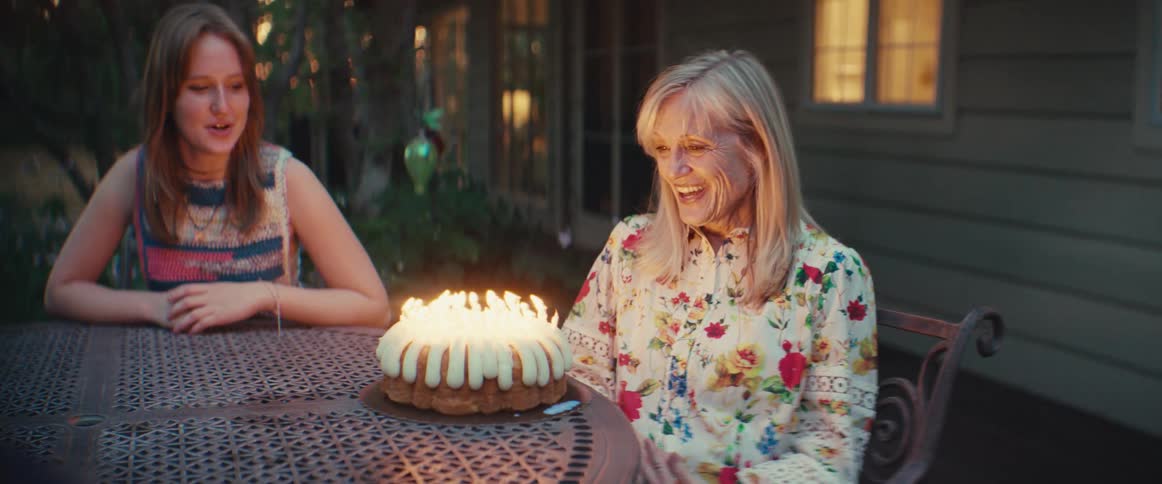 A woman sitting at a table outside at night blows out the candles on a cake