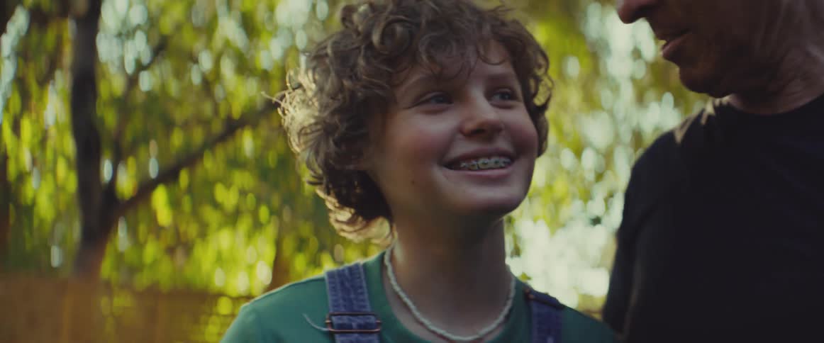 A father hugs his teenage son outdoors under the trees in the woods at midday both smiling warmly