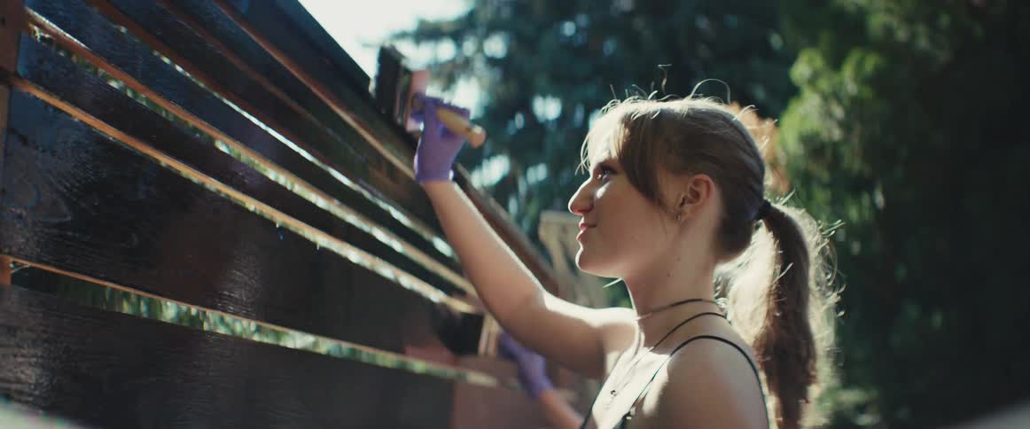 A young teenage girl uses a brush to paint a wooden fence wall surrounded by trees on a sunny day