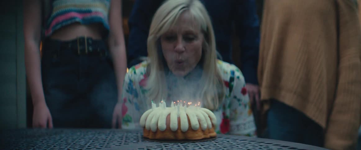 A smiling blonde woman in a white floral shirt sits at a dark outdoor table with a bundt cake topped with lit candles in front of her surrounded by three other people partially visible in the background