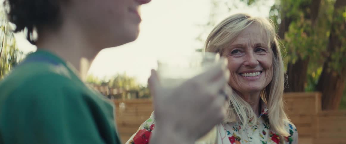 A closeup shows an older woman with long blonde hair smiling genuinely as she holds a glass and listens to someone offcamera the scene is outdoors and brightly lit with sunlight filtering through the trees suggesting a pleasant social gathering