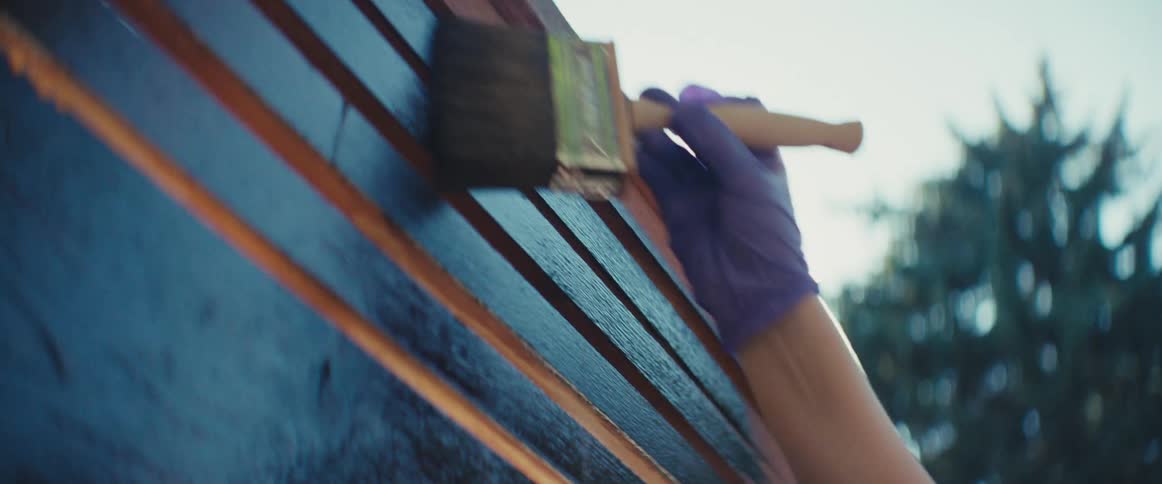 Woman with tiedback hair and sleeveless top reaches wooden structure wearing purple gloves in sunny outdoor setting