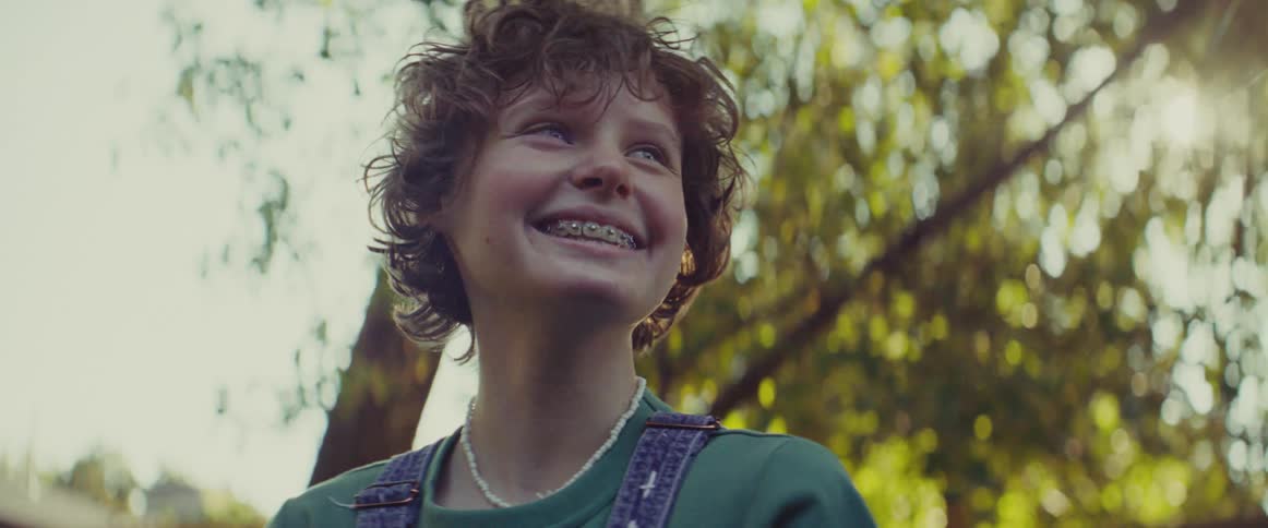 The video is a lowangle closeup shot of a young person with curly brown hair and braces smiling brightly toward the skythey are wearing dark green overalls over a tshirt and standing outdoors beneath the sundappled green leaves of trees