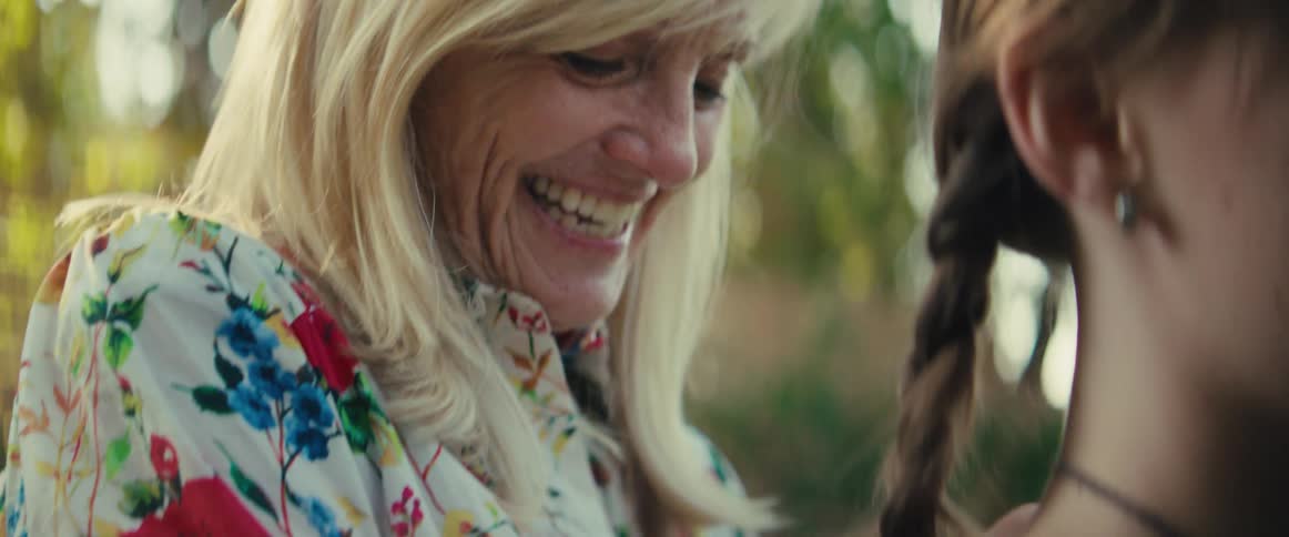 A woman with blonde hair smiles and laughs while interacting with a teenage girl braiding her hair and she wears a white shirt with a floral pattern both are in an outdoor setting