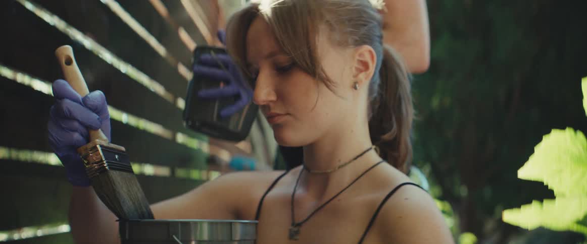 A close view shows a woman in a black top mixing paint in a metal can with a paintbrush while another persons hand wearing a purple glove paints a wooden slatted fence in a bright outdoor setting
