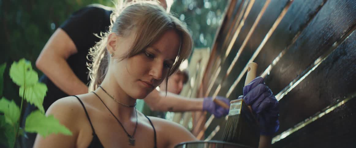 The video features a closeup of a young woman who is painting or staining a wooden fence outdoorsshe is concentrating on her work holding a paintbrush in one hand wearing a purple glove with another person also visible in the background painti