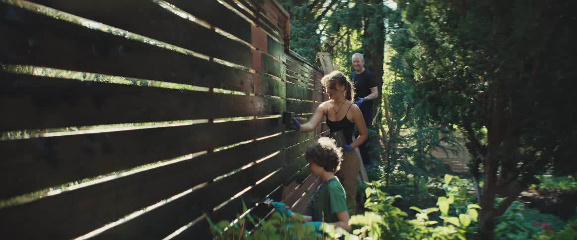 A woman a child and a man on a ladder paint a slatted wooden fence in a sunlit garden