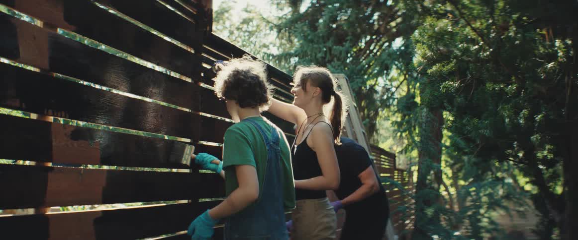 A family paints a wooden fence together in their backyard on a sunny day