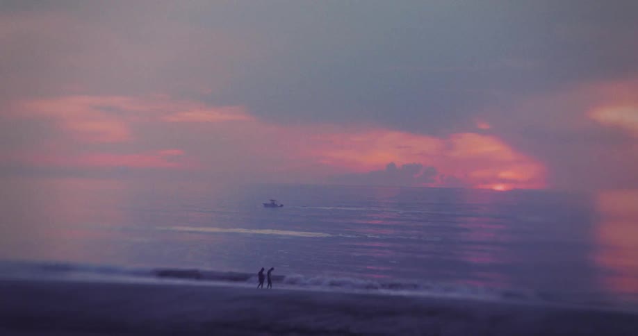 Walkers observe a distant boat under a pink and violet sunset