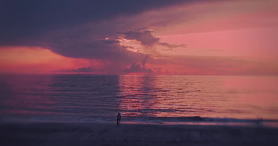 A lone figure stands on a beach facing the water under a vast dramatic sky dominated by heavy deep pink and purple clouds reflecting in the ocean the person is silhouetted against the bright horizon creating a sense of solitude and vastness
