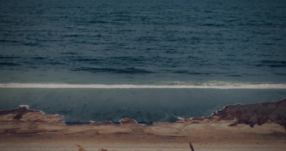 The video is a static shot of a wide moody ocean horizon with dark waves repeatedly crashing toward the shorethe foreground of the beach is a mix of wet and dry sand with the tide receding slightly after each incoming wave