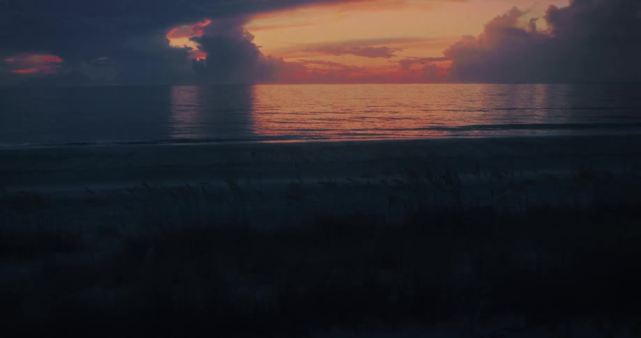 The video shows a sunrise or sunset over the ocean with the suns warm orange glow reflected on the waters surface dark heavy clouds frame the bright light above the horizon the foreground is a dark sandy beach with gentle waves rolling in