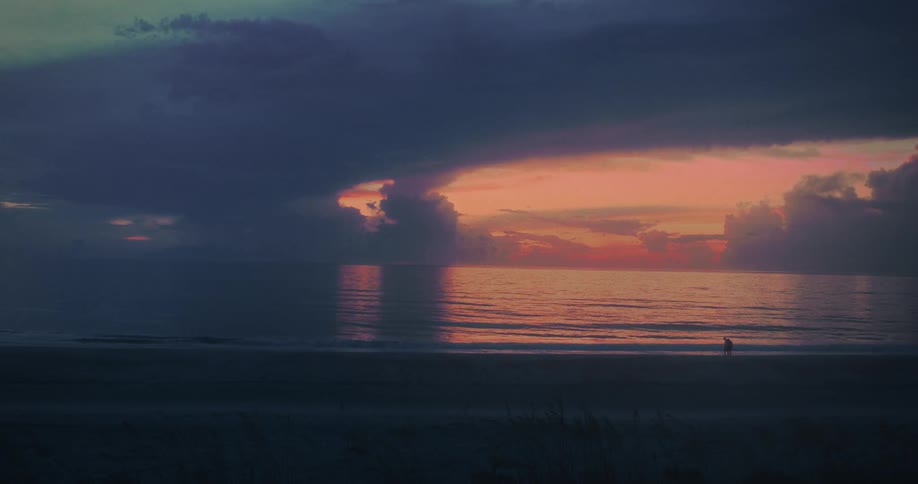 A couple walks along the wet sand of a vast ocean beach with small waves washing in under a sky with dark clouds and an intense orange sunset reflecting on the water