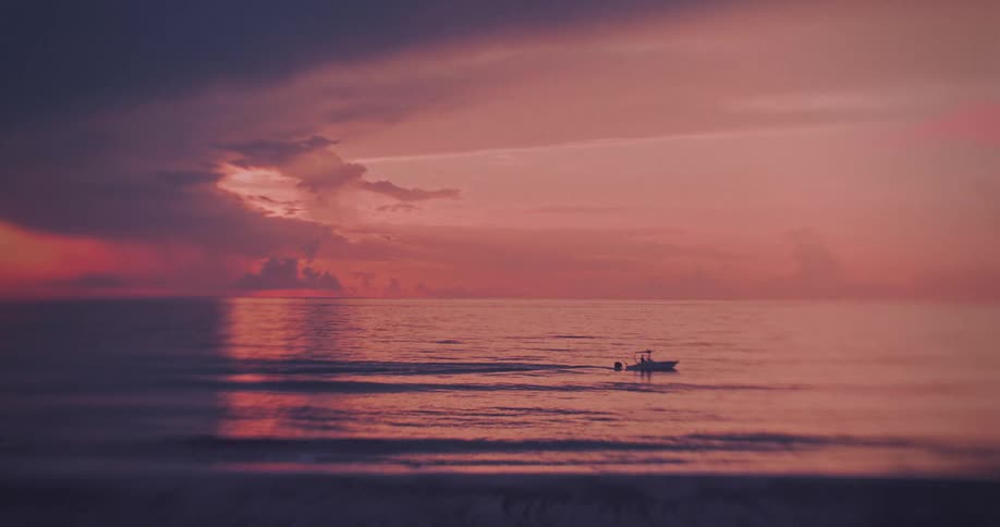 A small fishing boat motors slowly across the calm ocean under a vast dramatic twilight sky painted in shades of pink orange and deep purple the scene is characterized by a strong almost painterly color palette of the sunset reflecting on the dark r