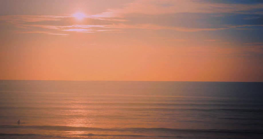 Waves roll into shore as the sun glows brightly through clouds in an orange sky