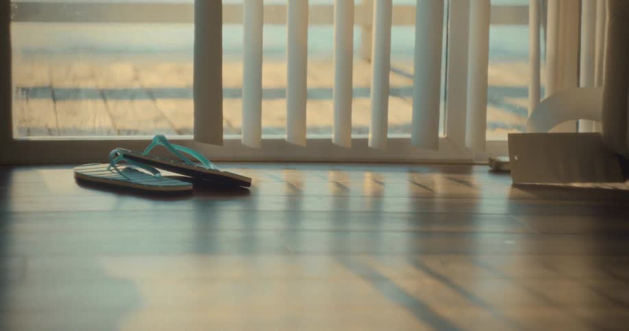 The scene is an interior shot of a room with polished wooden floors where a pair of blue and lightcolored flipflops rests near a sliding glass doorthe background shows vertical white blinds that are slightly open allowing bright golden sunlight t