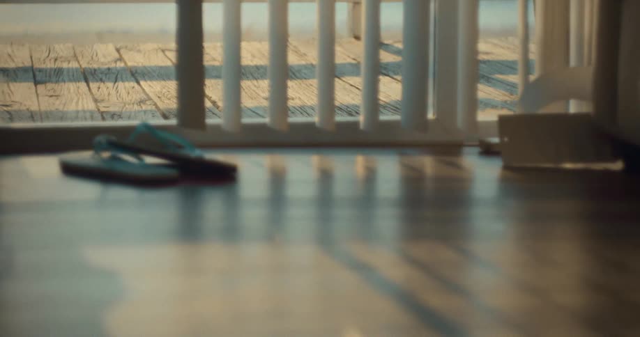 A low interior close view of wooden floor shows a pair of blue flip flops resting near a sunlit window with vertical blinds and a wooden pier visible outside