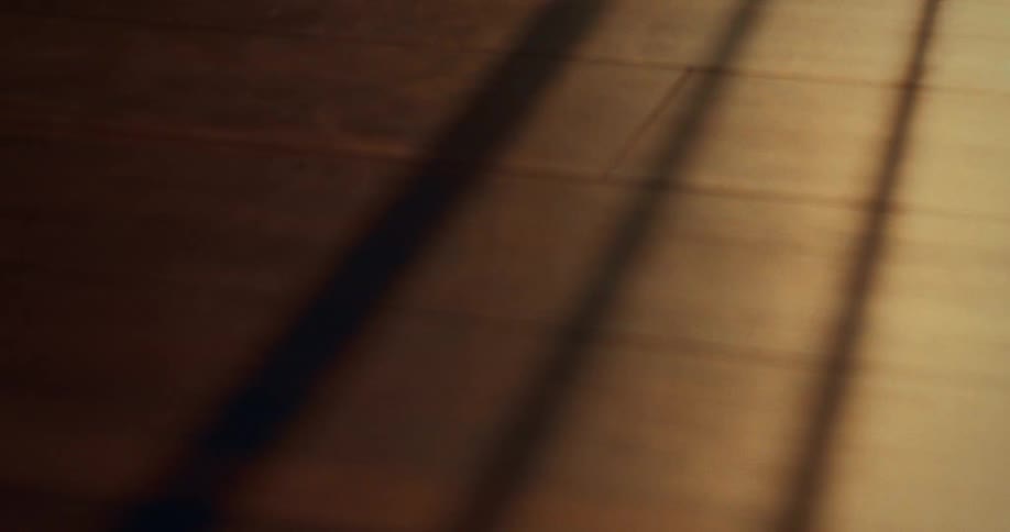 The video is a closeup shot of a wooden floor with a warm amber tone where light is cast through a window long dark shadows from an unseen object possibly window bars or a frame dramatically sweep across the floor and eventually cover the entire fr