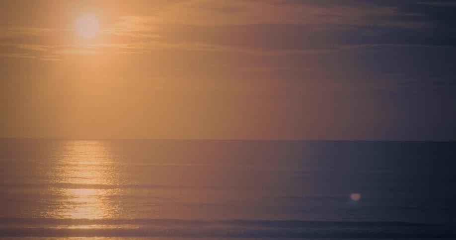 A bright sun hangs high in a golden orange sky above the ocean horizon casting a shimmering light path across the gently moving dark blue water