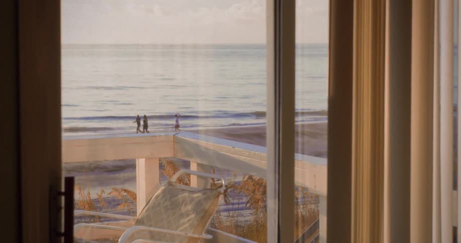 A beach view through sliding doors shows a balcony chair and a few people walking along the shoreline