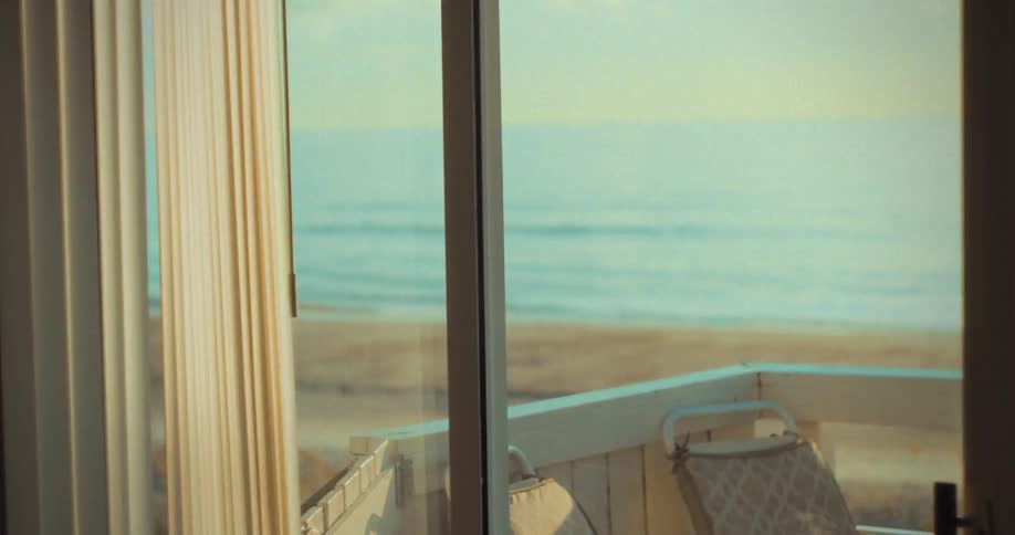 A sunlit ocean view is seen through sliding glass doors with a beachside balcony and two chairs in the foreground