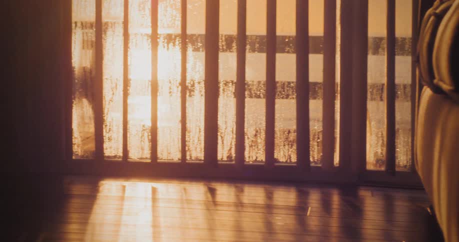 Golden bright sunlight streams through a slatted window covered with condensation or rain onto a dark wooden floor in an otherwise dim room with a soft lens flare effect
