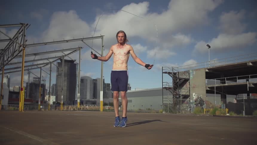 A young man with long, blonde hair jumping rope in an industrial area under a cloudy sky