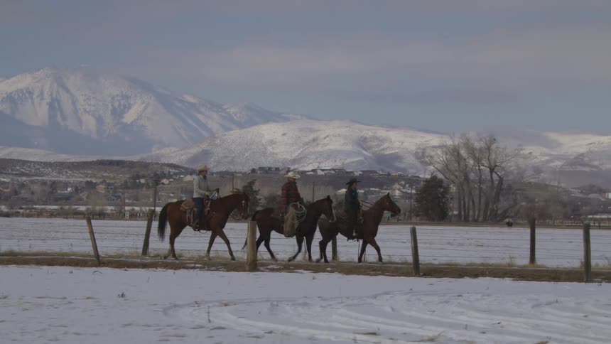 Three people on three horses walk along a fence line in a snowcovered field with snowcapped mountains rising prominently in the background under a pale sky