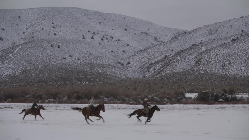 Three people ride horses across a flat area of white ground the horses gallop from left to right and large hills covered in snow and dark brush rise steeply in the background under a gray sky