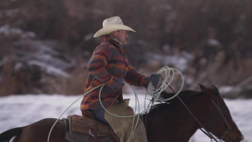 A cowboy wearing a white hat plaid shirt and gloves gallops on a brown horse holding a coiled lasso rope in a snowy outdoor field