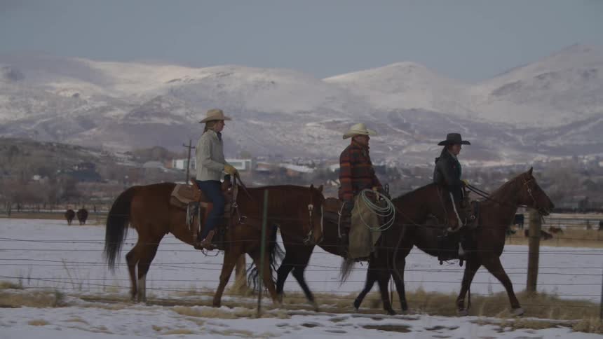 Three people wearing cowboy hats ride horses in a line from left to right across a partially snowcovered field a wire fence runs horizontally across the foreground and large snowdusted mountains dominate the background