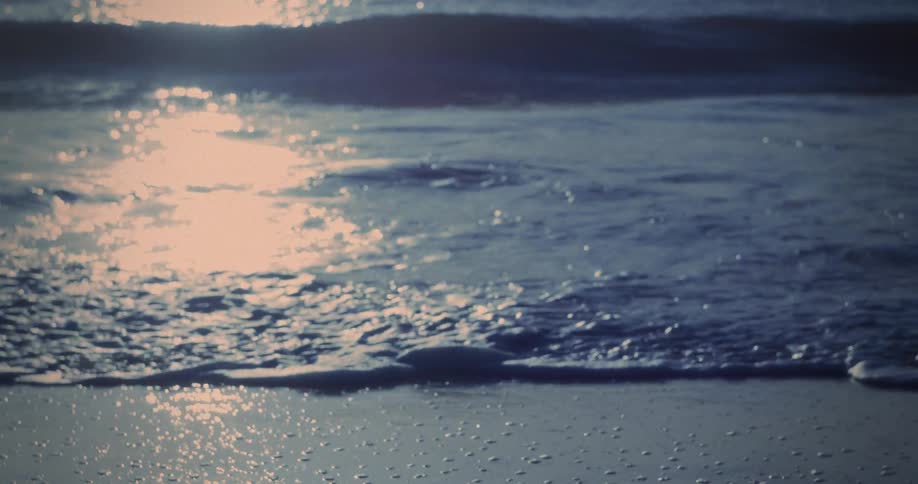 Sunlight reflects brightly on the surface of the water and wet sand ocean waves roll in and break near the bottom of the frame and the scene is blurred and dark above the horizon