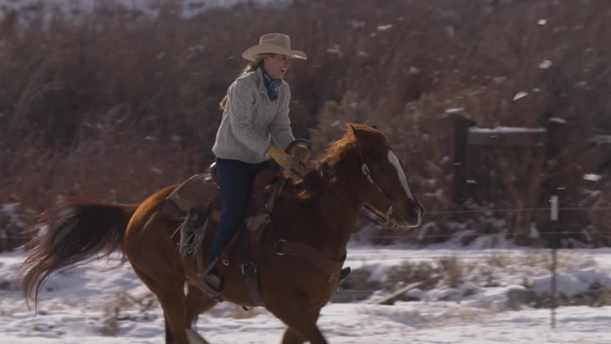 Cowgirl wearing a hat riding a horse through the snow