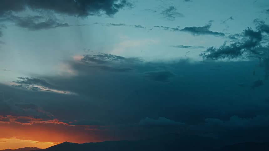 A dramatic wide shot captures a stormy twilight sky with heavy bluegray clouds dominating the frame as they slowly move in the lower left a sliver of intense orange light from the setting or rising sun is visible above a dark mountain range